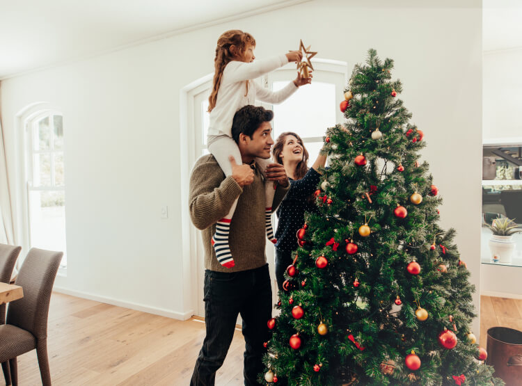 Little girl sitting on dad's shoulders adding the start to the holiday tree while mom watches